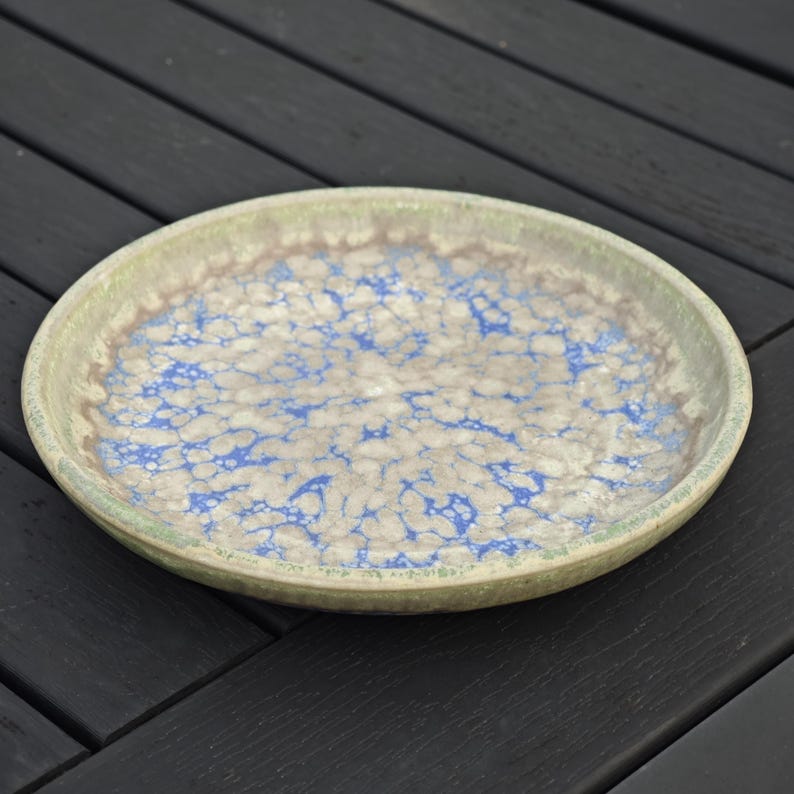 May include: A round ceramic plate with a speckled blue and white pattern on a beige background. The rim is a muted green. The plate is likely handmade and suitable for serving food.