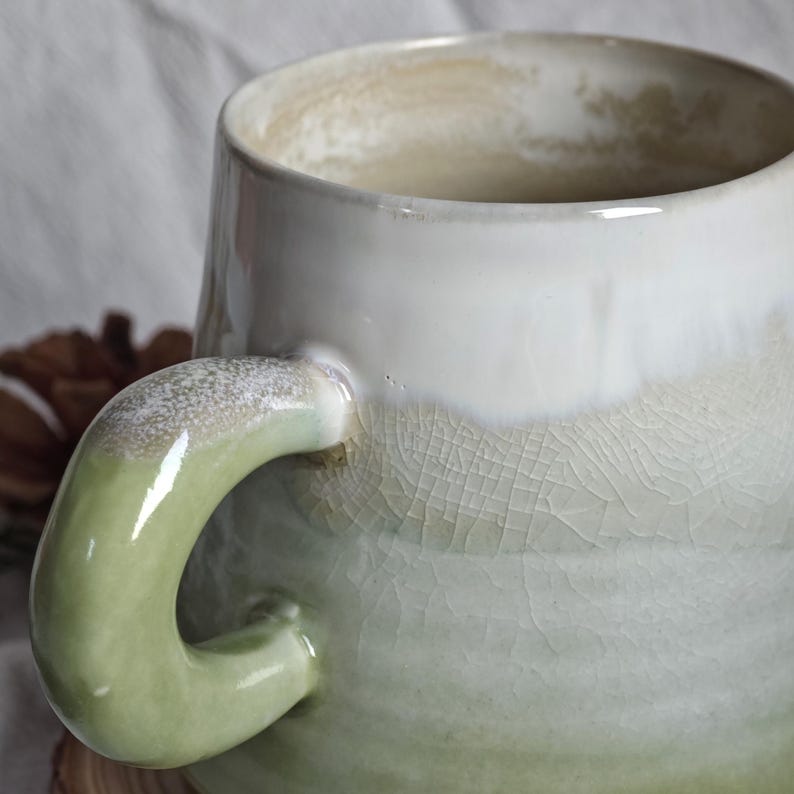 May include: A handmade ceramic mug with a unique, crackled glaze. The mug features a rounded body with a wide opening and a curved handle. The color palette includes shades of white, green, and beige, creating a rustic and artistic aesthetic.