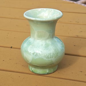 Puede incluir: Un jarr&oacute;n de cer&aacute;mica verde claro con cuerpo bulboso y borde acampanado. El jarr&oacute;n tiene un patr&oacute;n de esmalte cristalino con detalles en blanco y verde claro. El jarr&oacute;n mide aproximadamente 15 cm de alto.
