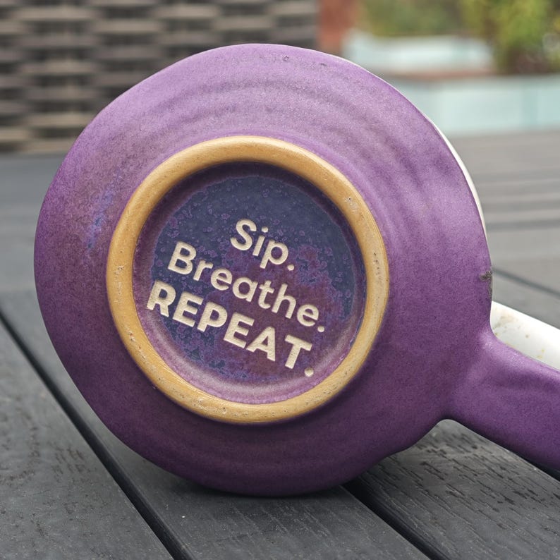Op de afbeelding: Een paarse keramische mok met een wit handvat, van onderaf gezien. De bodem toont de witte tekst "Sip. Breathe. REPEAT." omringd door een beige ring. De mok staat op een donkergrijze houten ondergrond.