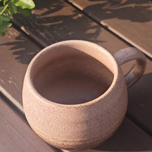 Może przedstawiać: Okrągły, nakrapiany r&oacute;żowy kubek ceramiczny z solidnym uchwytem. Na zewnętrznej stronie znajduje się subtelny, wygrawerowany rysunek liniowy kota w pobliżu podstawy. Kubek ma teksturowane, matowe wykończenie z ciemniejszym, ziemistym odcieniem.