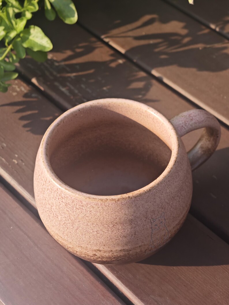 Może przedstawiać: Okrągły, nakrapiany r&oacute;żowy kubek ceramiczny z solidnym uchwytem. Na zewnętrznej stronie znajduje się subtelny, wygrawerowany rysunek liniowy kota w pobliżu podstawy. Kubek ma teksturowane, matowe wykończenie z ciemniejszym, ziemistym odcieniem.