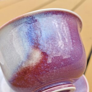 May include: A ceramic teacup with a glossy, iridescent finish. The cup transitions from a deep burgundy at the base to shades of blue and white near the rim. The cup sits on a matching saucer, creating a cohesive set.