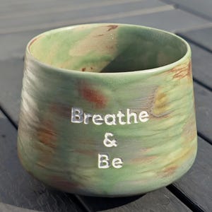 May include: A green and brown ceramic cup with the words "Breathe & Be" etched into the side. The cup has a wide opening and a rounded shape. The glaze has a marbled effect.