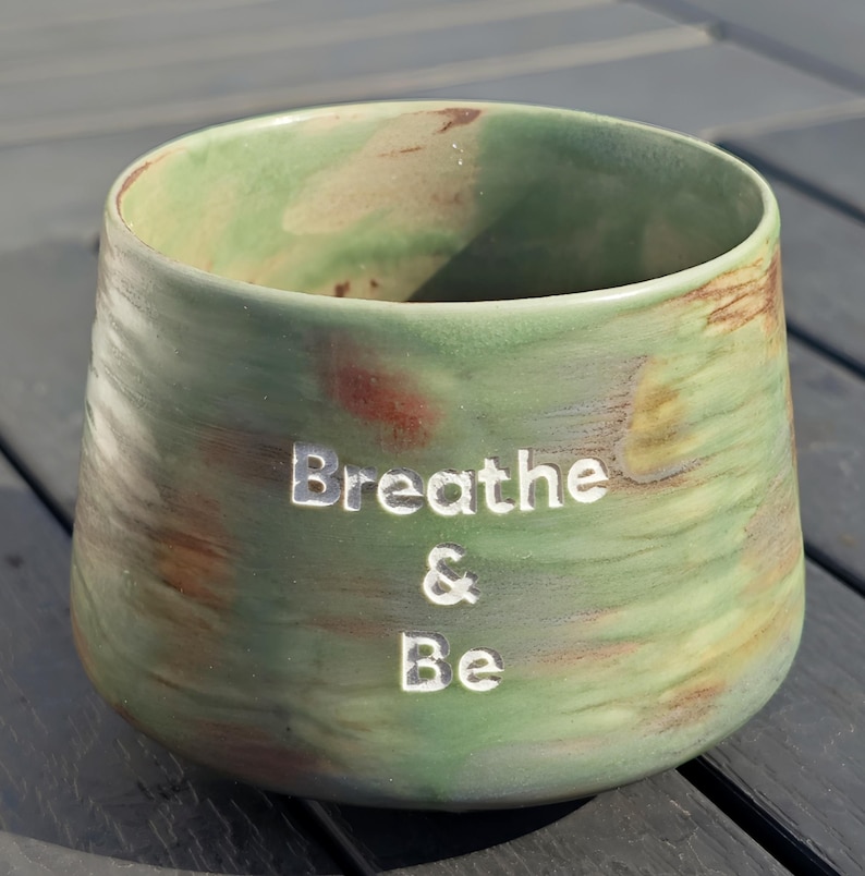 May include: A green and brown ceramic cup with the words "Breathe & Be" etched into the side. The cup has a wide opening and a rounded shape. The glaze has a marbled effect.