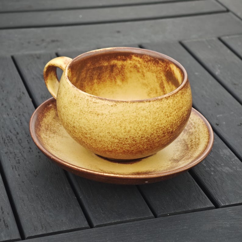 May include: A round, yellow-brown ceramic teacup and saucer set. The cup has a rounded shape with a handle, and the saucer is circular. The set has a speckled texture and a brown rim. The teacup and saucer are on a dark wooden surface.