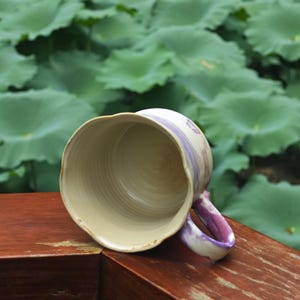 May include: A handmade ceramic mug with a cream-colored interior and a wavy rim. The exterior features a light purple and white painted design. The handle is also painted with purple and white. The mug is sitting on a wooden surface.
