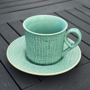 May include: A teal ceramic teacup and saucer set. The cup has a rounded handle and a textured woven pattern. The saucer is round with a matching pattern and a cream-colored rim. The set is on a dark wooden surface.