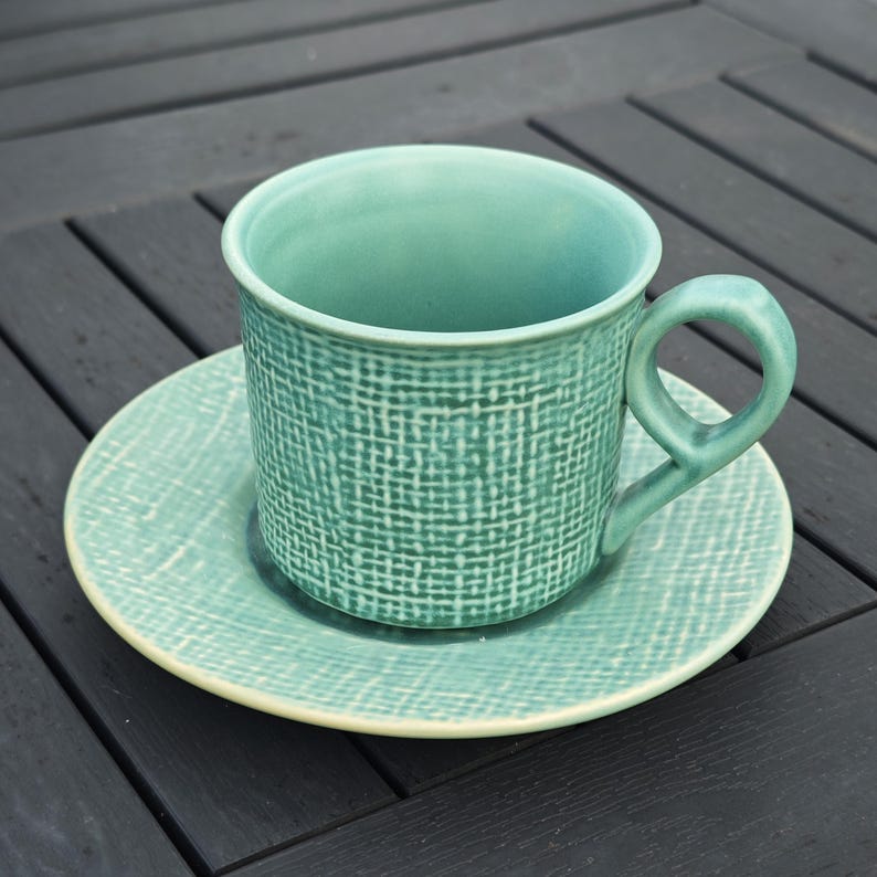 May include: A teal ceramic teacup and saucer set. The cup has a rounded handle and a textured woven pattern. The saucer is round with a matching pattern and a cream-colored rim. The set is on a dark wooden surface.