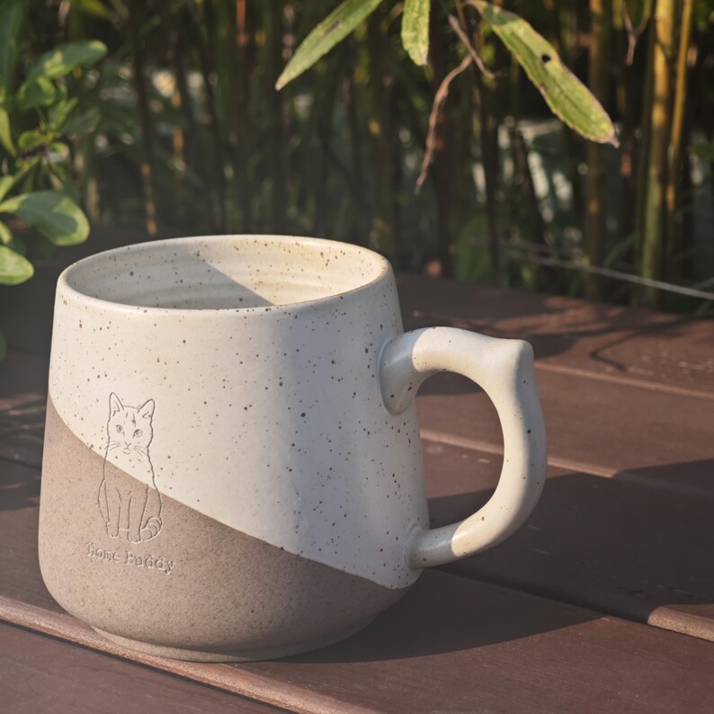 May include: A ceramic mug with a two-tone design featuring a speckled cream upper section and a solid matte brown lower section. A line drawing of a cat is centred on the front with the text "rose buddy" printed below the cat illustration.