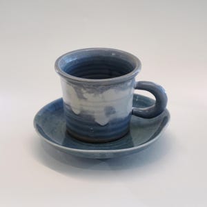 May include: A blue and white ceramic mug and saucer set. The mug has a rounded handle and a cylindrical shape. The saucer is round and complements the mug. The set is glazed with a marbled effect. Ideal for serving tea or coffee, the set is a handcrafted item.