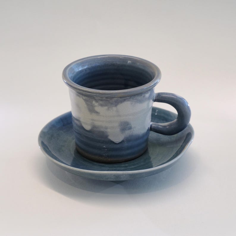 May include: A blue and white ceramic mug and saucer set. The mug has a rounded handle and a cylindrical shape. The saucer is round and complements the mug. The set is glazed with a marbled effect. Ideal for serving tea or coffee, the set is a handcrafted item.