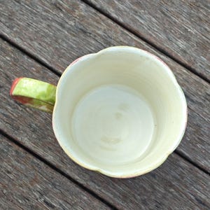 May include: A handmade ceramic mug with a unique, scalloped rim. The interior is a creamy off-white, contrasting with the green and red handle. The mug sits on a wooden surface, showcasing its handcrafted details.
