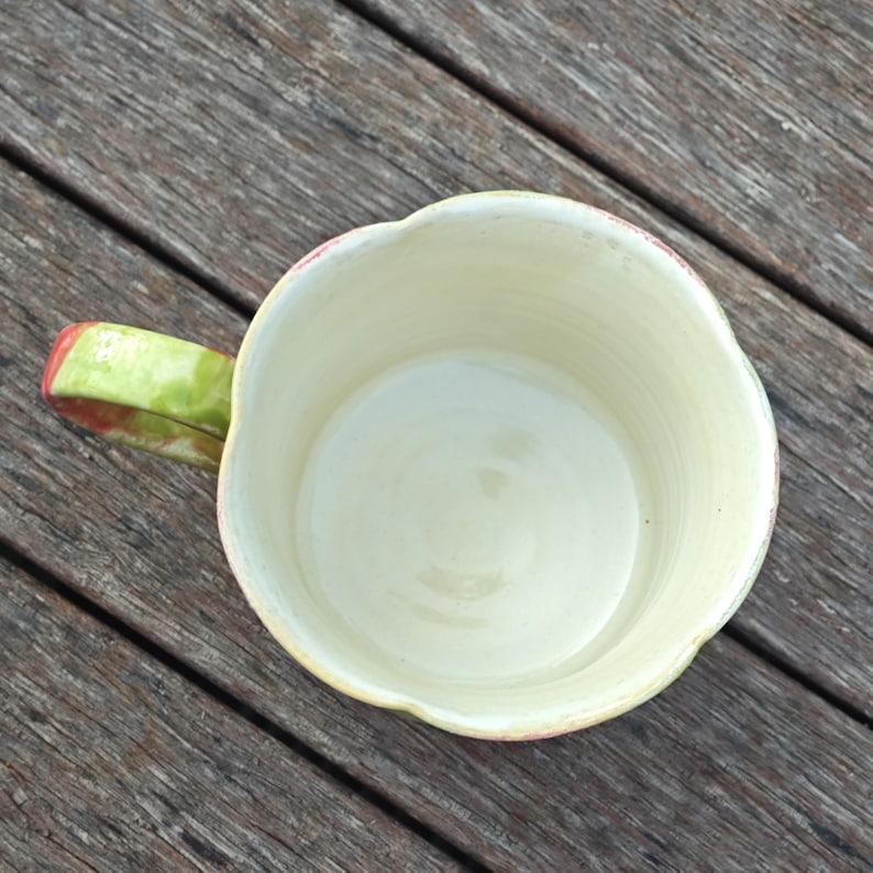 May include: A handmade ceramic mug with a unique, scalloped rim. The interior is a creamy off-white, contrasting with the green and red handle. The mug sits on a wooden surface, showcasing its handcrafted details.