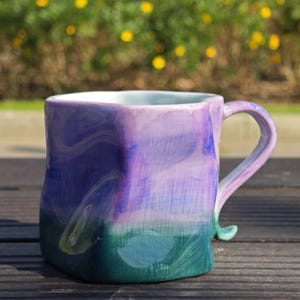 May include: Hand-painted ceramic mug with a unique, square shape. The mug features a gradient of colors, transitioning from deep green at the base to shades of blue and purple at the top. The handle is a matching color and design.