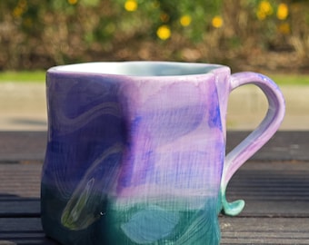 May include: Hand-painted ceramic mug with a unique, square shape. The mug features a gradient of colors, transitioning from deep green at the base to shades of blue and purple at the top. The handle is a matching color and design.