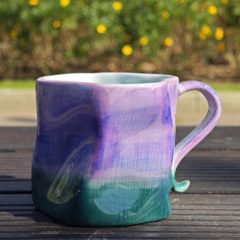 May include: Hand-painted ceramic mug with a unique, square shape. The mug features a gradient of colors, transitioning from deep green at the base to shades of blue and purple at the top. The handle is a matching color and design.