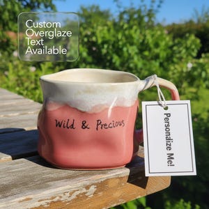 May include: A handmade ceramic mug with a pink and white glaze. The mug has the words "Wild & Precious" written on it. A tag that says "Personalize Me!" is attached to the handle. The mug is sitting on a wooden surface.