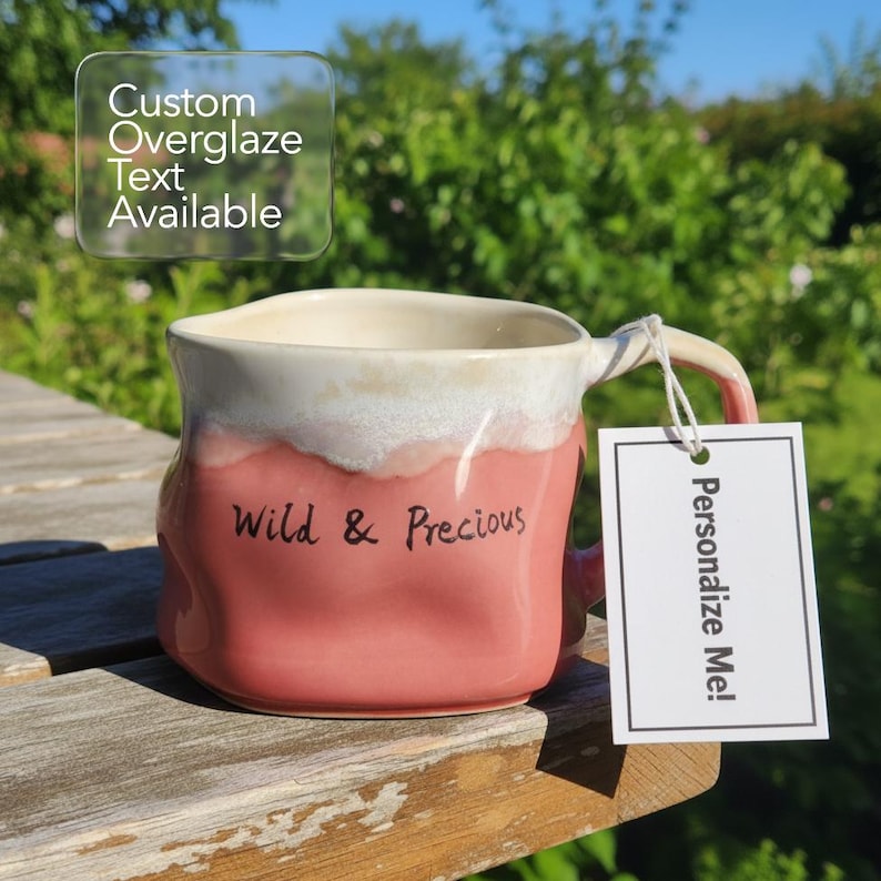 May include: A handmade ceramic mug with a pink and white glaze. The mug has the words "Wild & Precious" written on it. A tag that says "Personalize Me!" is attached to the handle. The mug is sitting on a wooden surface.