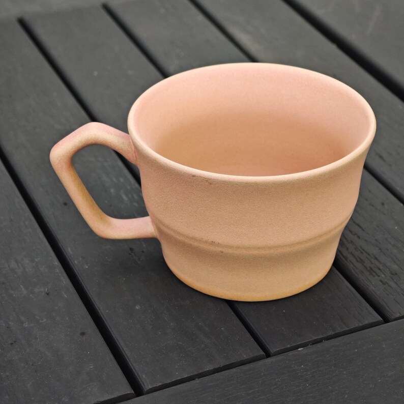May include: A peach-colored ceramic mug with a rounded body and a handle. The mug has a textured surface and a simple, classic design. The mug is sitting on a dark gray wooden surface.