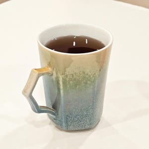 May include: A ceramic mug with a unique geometric handle, filled with a dark liquid. The mug features a gradient of colors, transitioning from beige to green and ending in a speckled blue at the base. The interior is white.