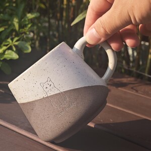 May include: A two-toned ceramic mug with a speckled cream upper section and a solid dark grey lower section. The mug features a line art illustration of a cat and the text Home Buddy etched into the surface, complete with a curved handle.