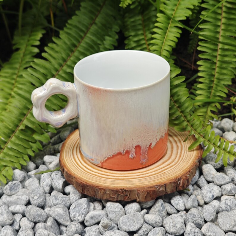 May include: A handmade ceramic mug with a unique flower-shaped handle. The mug features a white glaze that transitions to a peach color at the base. It sits on a small wooden coaster, surrounded by gray stones and green foliage.