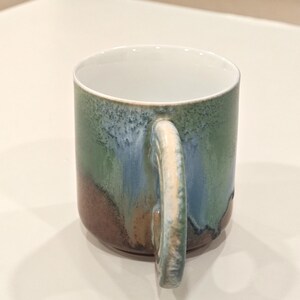 K&ouml;nnte beinhalten: Eine Keramiktasse mit einzigartiger Glasur. Die Tasse hat einen wei&szlig;en Innenraum und einen Henkel. Die Au&szlig;enseite weist eine Mischung aus Farben auf, darunter Gr&uuml;n, Blau und Braun. Die Tasse ist f&uuml;r hei&szlig;e oder kalte Getr&auml;nke geeignet.