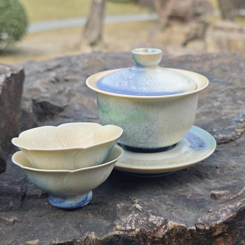 May include: A ceramic tea set with a lidded gaiwan, a saucer, and two small teacups. The gaiwan and saucer have blue and green accents. The teacups are a light yellow color with a petal design.