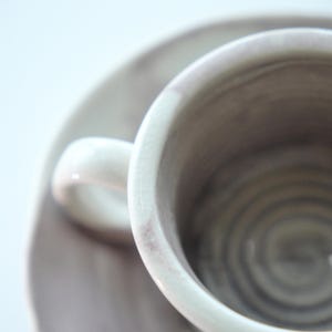 May include: Close-up of a ceramic teacup and saucer set. The cup and saucer are a muted gray color with subtle pinkish-purple accents. The cup has a small handle and sits on a matching saucer, creating a minimalist aesthetic.