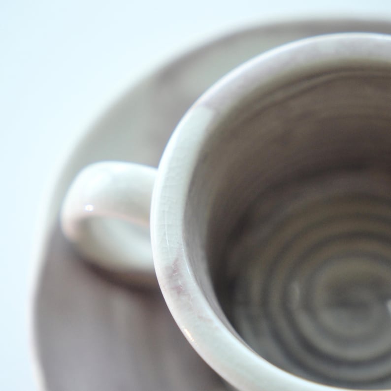 May include: Close-up of a ceramic teacup and saucer set. The cup and saucer are a muted gray color with subtle pinkish-purple accents. The cup has a small handle and sits on a matching saucer, creating a minimalist aesthetic.