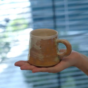 May include: A handmade ceramic mug with a rounded handle. The mug has a light brown exterior with a white rim and interior. The mug is held in a person's hand.