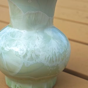 Puede incluir: Un jarr&oacute;n de cer&aacute;mica verde p&aacute;lido con un cuerpo bulboso y un cuello acampanado. El jarr&oacute;n presenta un patr&oacute;n cristalino &uacute;nico en blanco, creando un dise&ntilde;o texturizado y org&aacute;nico. Es un objeto decorativo.
