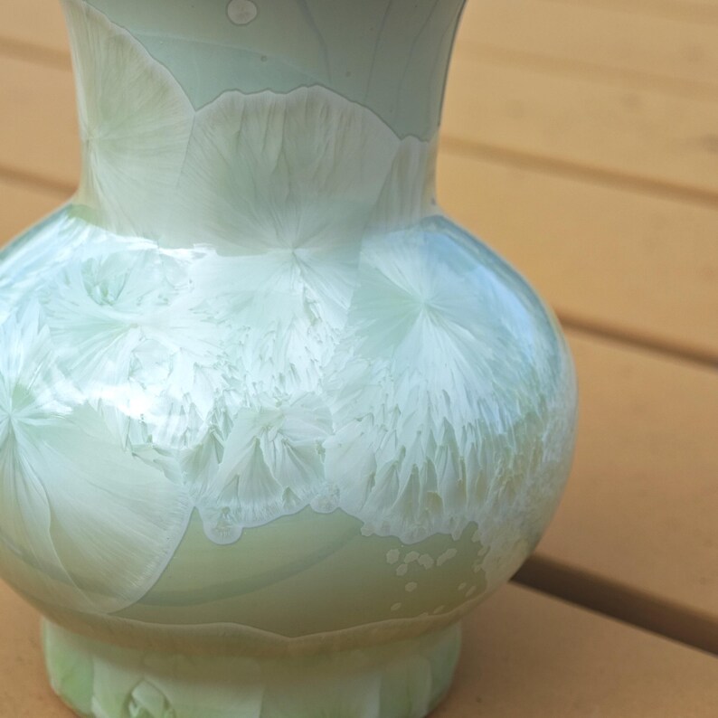 Puede incluir: Un jarr&oacute;n de cer&aacute;mica verde p&aacute;lido con un cuerpo bulboso y un cuello acampanado. El jarr&oacute;n presenta un patr&oacute;n cristalino &uacute;nico en blanco, creando un dise&ntilde;o texturizado y org&aacute;nico. Es un objeto decorativo.
