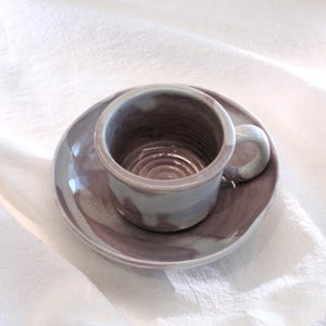 May include: A handmade ceramic teacup and saucer set. The cup and saucer are a muted purple color with a white handle. The cup has a circular design on the inside. The set is on a white cloth.