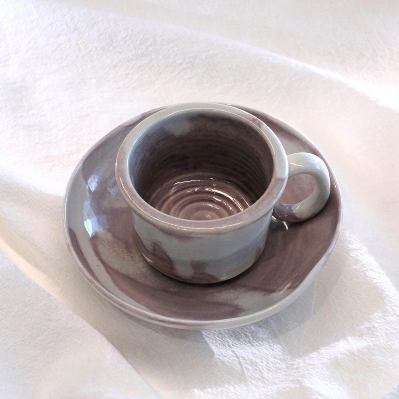 May include: A handmade ceramic teacup and saucer set. The cup and saucer are a muted purple color with a white handle. The cup has a circular design on the inside. The set is on a white cloth.
