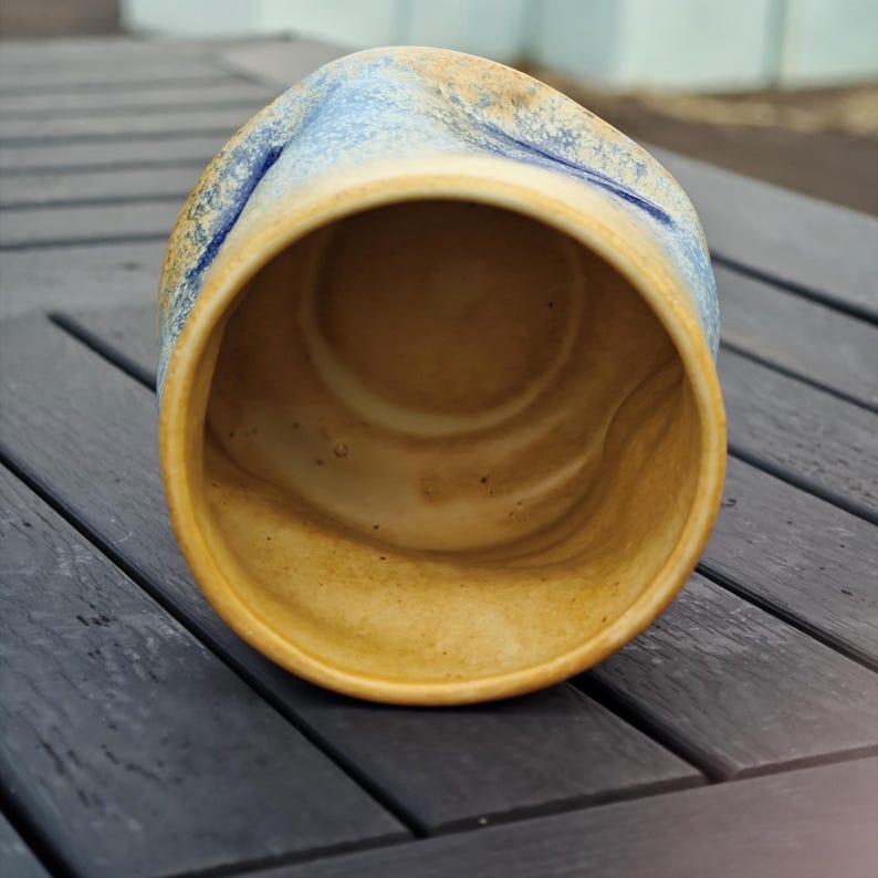 Peut inclure: Une tasse en c&eacute;ramique faite &agrave; la main avec un design textur&eacute; unique. La tasse pr&eacute;sente un m&eacute;lange de couleurs, notamment des nuances de jaune, de bleu et de marron. L'int&eacute;rieur est d'une teinte chaude et terreuse, et l'ext&eacute;rieur a une forme l&eacute;g&egrave;rement irr&eacute;guli&egrave;re.