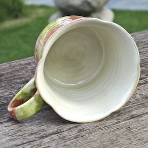 May include: A handmade ceramic mug with a wide, scalloped rim. The exterior features a blend of green and red hues, while the interior is a creamy white. The handle is also painted with green and red. The mug is set on a wooden surface.