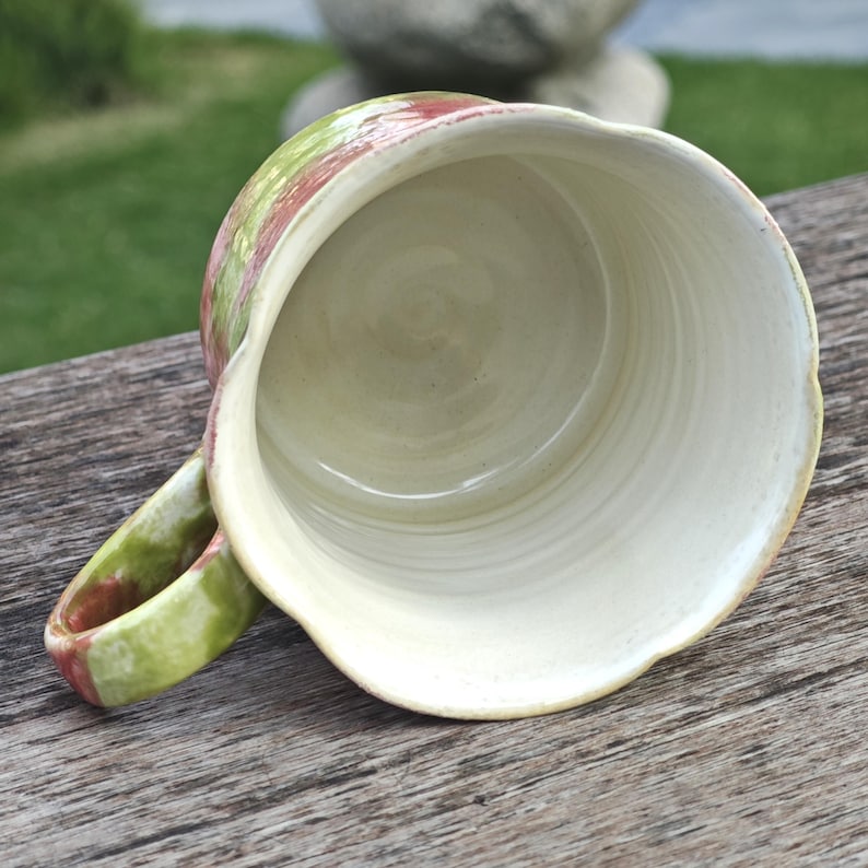 May include: A handmade ceramic mug with a wide, scalloped rim. The exterior features a blend of green and red hues, while the interior is a creamy white. The handle is also painted with green and red. The mug is set on a wooden surface.