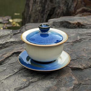 May include: A ceramic gaiwan tea set consisting of a bowl, a lid, and a saucer. The bowl is off-white, while the lid and saucer feature a deep blue glaze with a speckled texture. The lid has a small, rounded knob handle at the very top center.