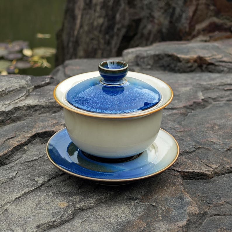 May include: A ceramic gaiwan tea set consisting of a bowl, a lid, and a saucer. The bowl is off-white, while the lid and saucer feature a deep blue glaze with a speckled texture. The lid has a small, rounded knob handle at the very top center.