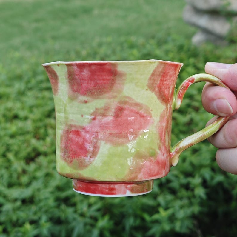 May include: Handmade ceramic mug with a unique design. The mug features a light green base with red splotches, a matching handle, and a white rim. The mug is held by a person's hand against a blurred green background.