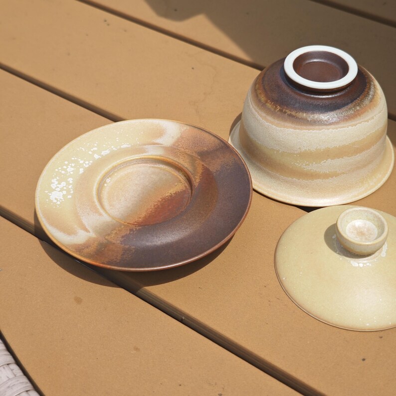 May include: A ceramic tea set consisting of a saucer, an inverted bowl, and a separate lid. The pieces feature a gradient glaze transitioning from creamy beige to deep brown, with a textured, speckled finish visible on the saucer and bowl.