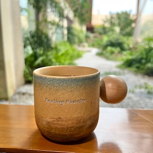 May include: A ceramic mug with a brown and blue gradient, featuring a wooden ball handle. The mug has the phrase "Feeling Peachy" printed on the front. The mug is sitting on a wooden surface.
