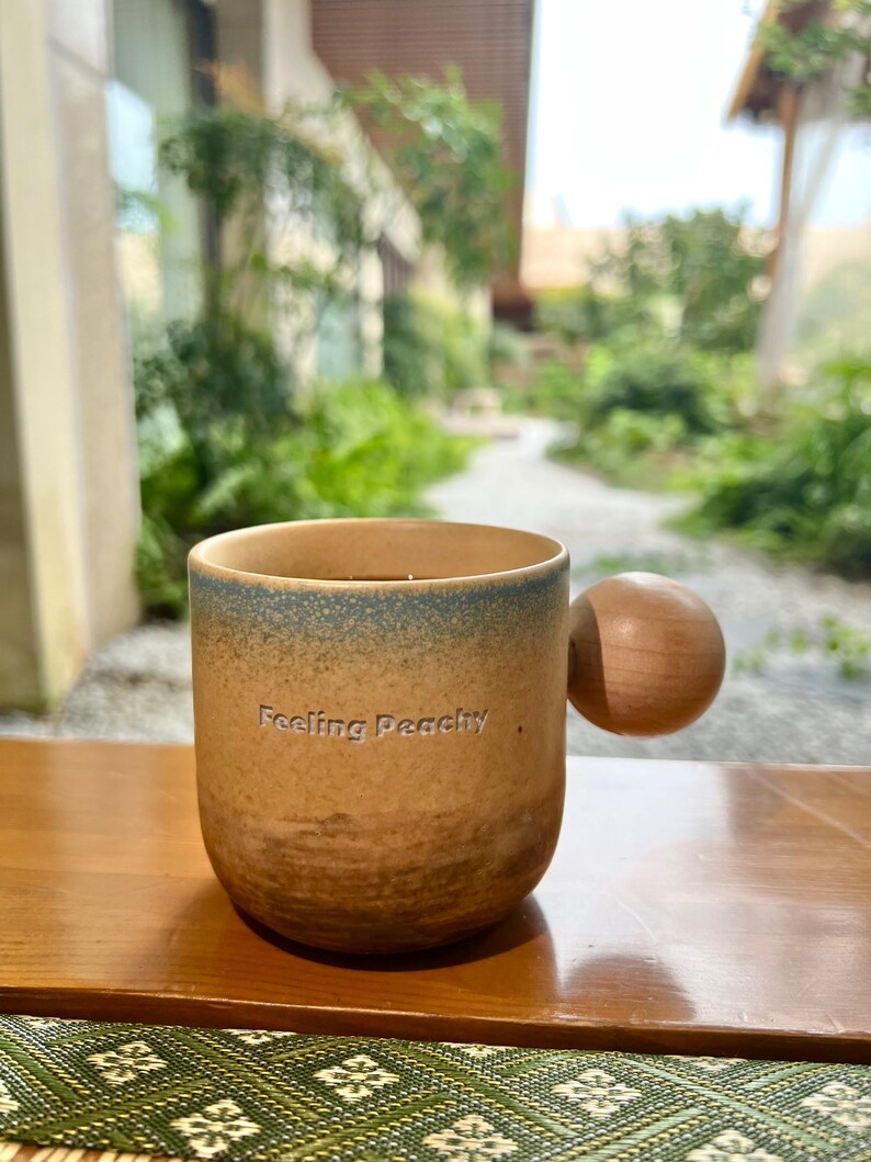 May include: A ceramic mug with a brown and blue gradient, featuring a wooden ball handle. The mug has the phrase "Feeling Peachy" printed on the front. The mug is sitting on a wooden surface.