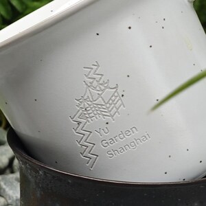 May include: Close-up of a white ceramic planter with a speckled finish. The planter features an embossed design of a pagoda and the text "Yu Garden Shanghai". The planter is stacked on top of a dark brown ceramic pot.