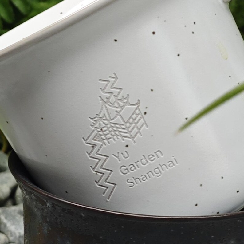 May include: Close-up of a white ceramic planter with a speckled finish. The planter features an embossed design of a pagoda and the text "Yu Garden Shanghai". The planter is stacked on top of a dark brown ceramic pot.