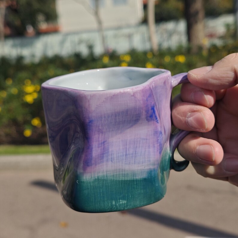 May include: A handmade ceramic mug with a unique, square shape and a handle. The mug features a gradient of colors, transitioning from teal at the base to purple and blue at the top. The interior is white.