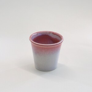 May include: A small, handmade ceramic cup with a gradient color scheme. The rim is a deep red, transitioning to a lighter pink and then a pale gray towards the base. The cup has a simple, cylindrical shape.