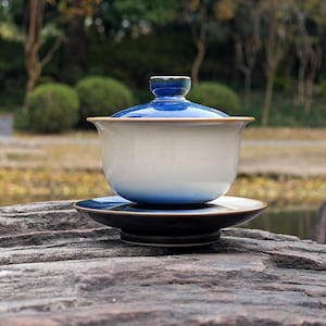 May include: A ceramic gaiwan tea set consisting of a bowl, a lid, and a saucer. The bowl features a gradient design transitioning from a deep blue at the base and rim to a creamy off-white center. The lid is deep blue, and the saucer is dark.